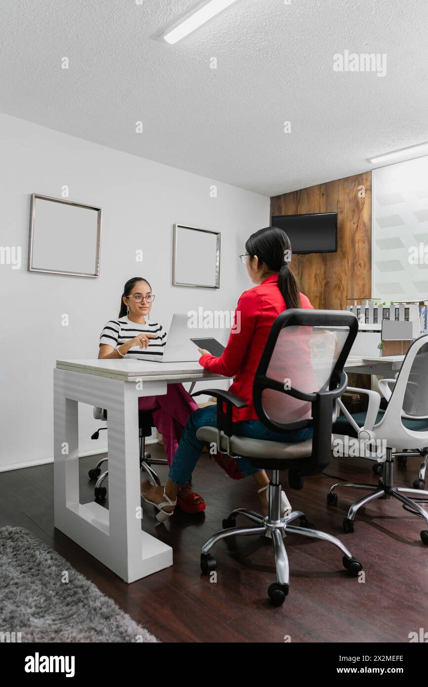 Deux femmes professionnelles latino-américaines qui s'engagent dans une conversation dans un bureau élégant et contemporain, illustrant le travail d'équipe et la communication d'entreprise. Banque D'Images