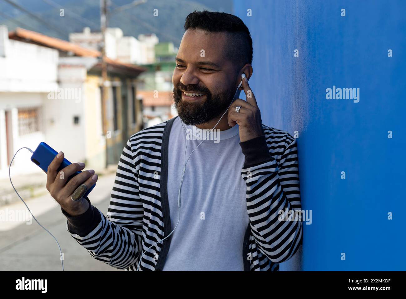 Mâle adulte (39 ans) barbe, moyen plat, utilise son téléphone portable avec un geste de bonheur et de positivisme. Habillé en pull rayé, t-shirt blanc, bleu Banque D'Images