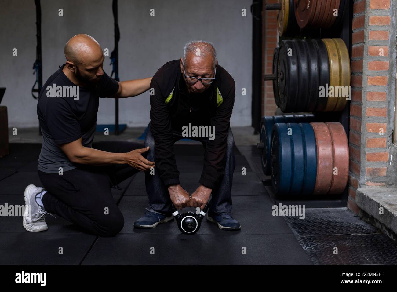Homme senior latino-américain avec son entraîneur personnel faire des exercices de rééducation et de thérapie tenant une kettlebell avec les deux mains dans la salle de gym. En bonne santé Banque D'Images