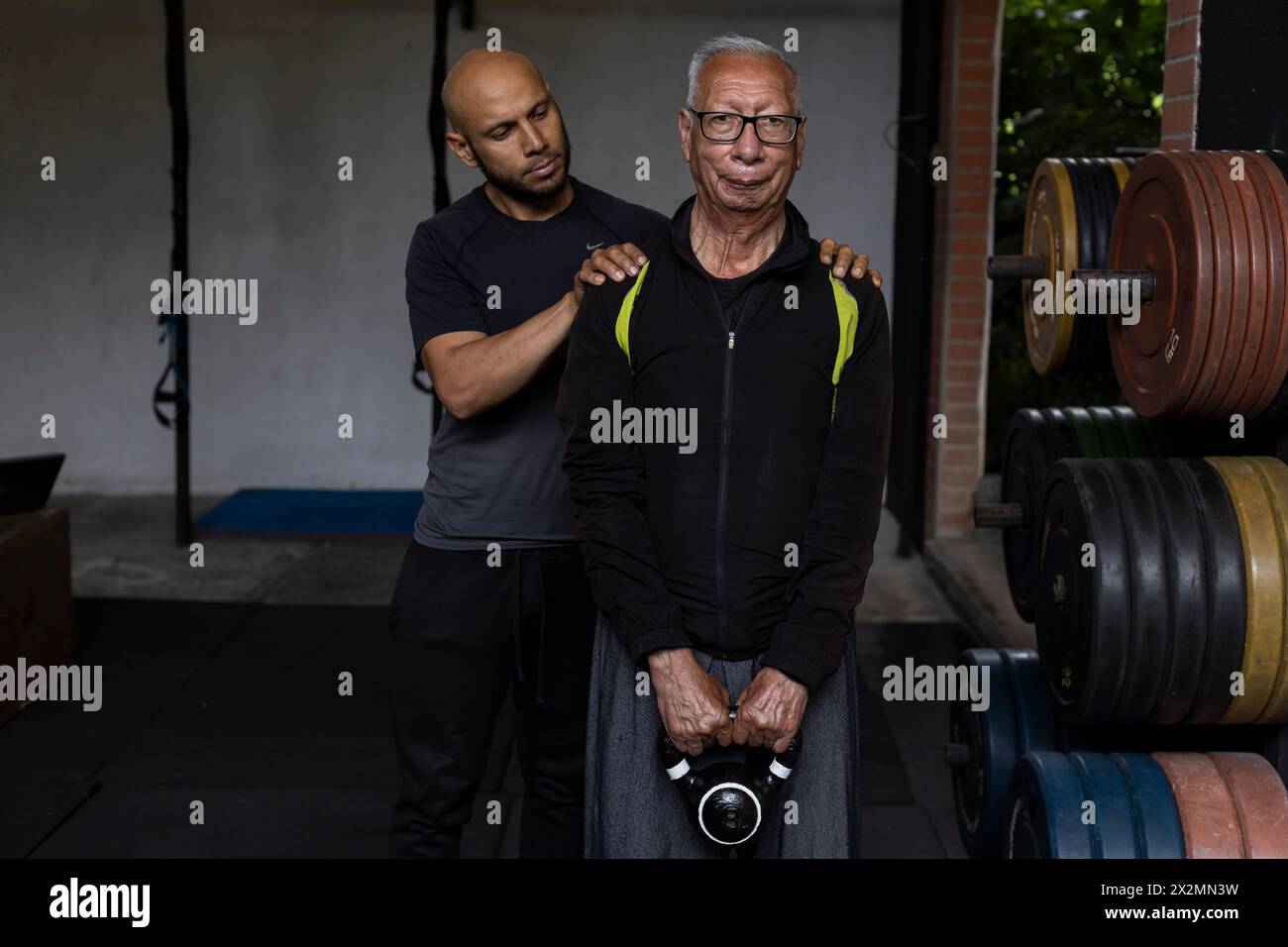 Homme senior latino-américain avec son entraîneur personnel faire des exercices de rééducation et de thérapie tenant une kettlebell avec les deux mains dans la salle de gym. En bonne santé Banque D'Images