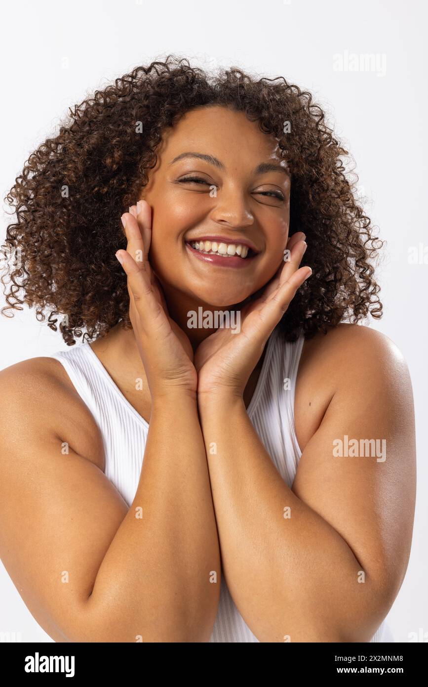 Modèle biracial grande taille avec des cheveux bruns bouclés souriant, touchant son visage sur blanc Banque D'Images