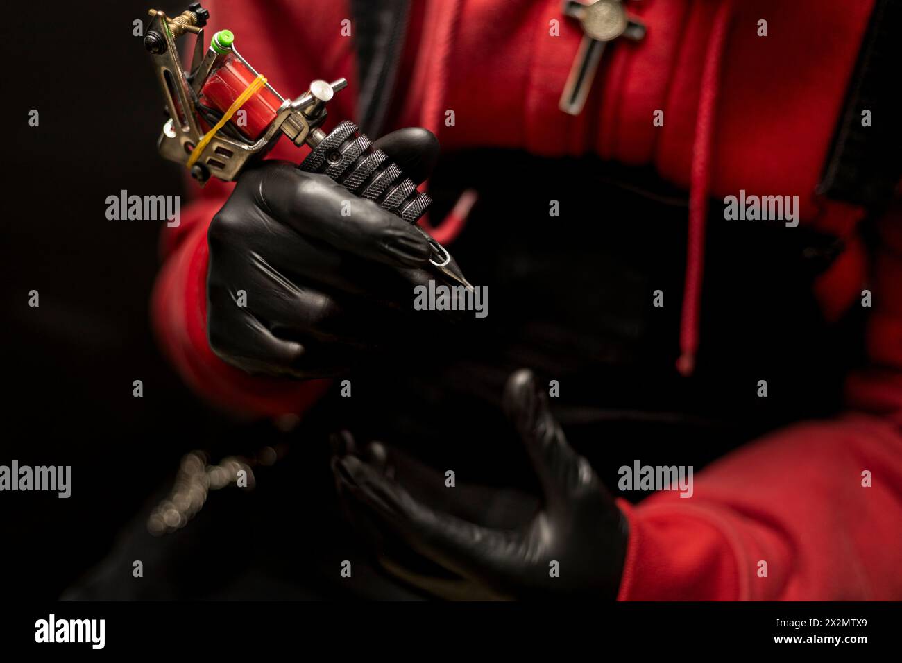 Mains de tatoueur montrant sa machine à tatouer. Il porte des gants noirs, un pull rouge et un fond noir. Concept d'art corporel Banque D'Images