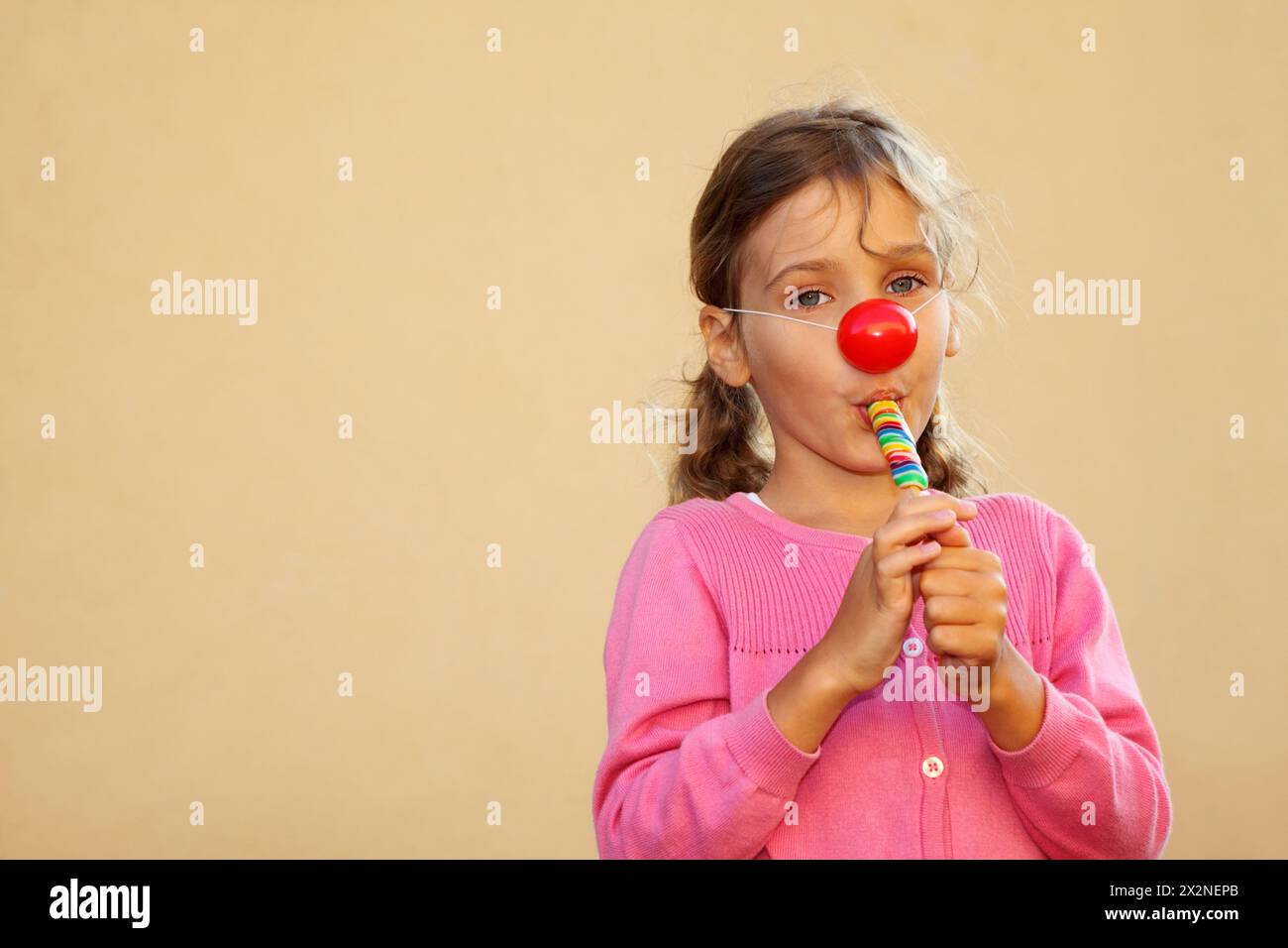 Fille portant un chemisier rose avec nez de clown rouge suce des bonbons multicolores. Banque D'Images