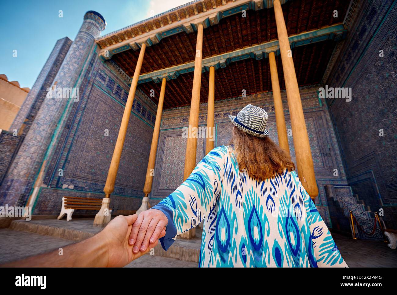 Femme touriste en robe ethnique à la cour intérieure de la salle de palais en mosaïque bleue avec colonne dirigeant l'homme tenu par la main à la ville antique Khiva en Ouzbékistan. Banque D'Images