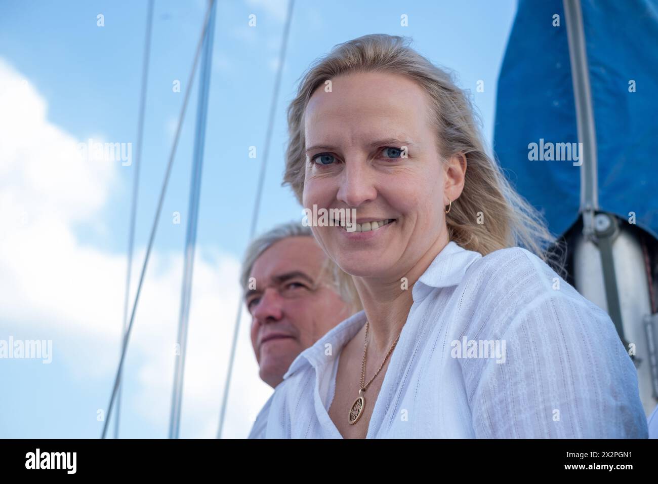 Femme souriante dans une blouse blanche avec un homme derrière elle sur un voilier, partageant un moment de joie sous un ciel bleu, idéal pour les loisirs et les thèmes de voyage. Bonjour Banque D'Images