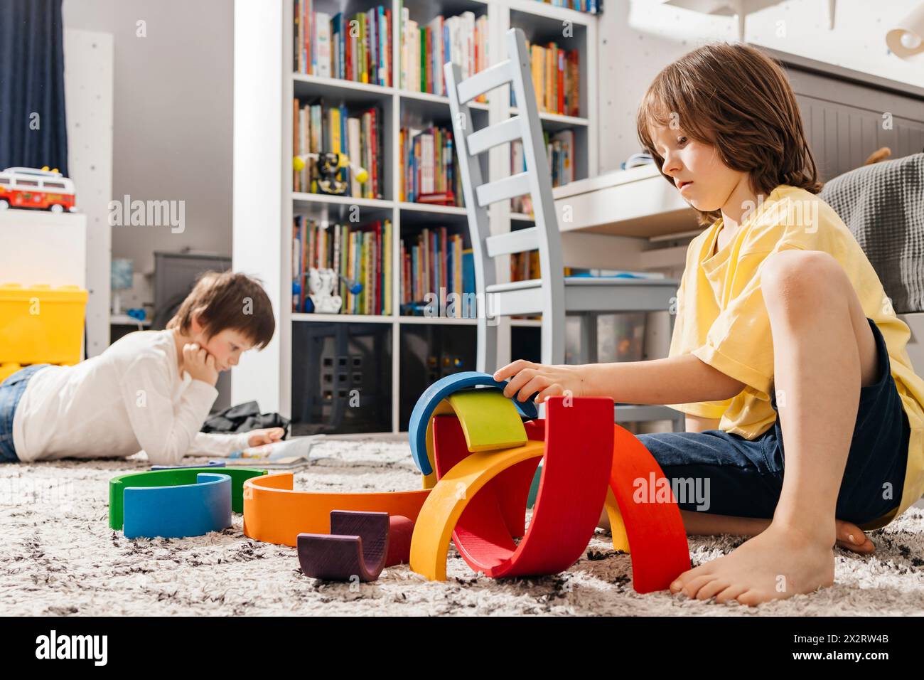 Garçon bâtissant pyramide avec des jouets colorés arc-en-ciel près du livre de lecture de frère Banque D'Images