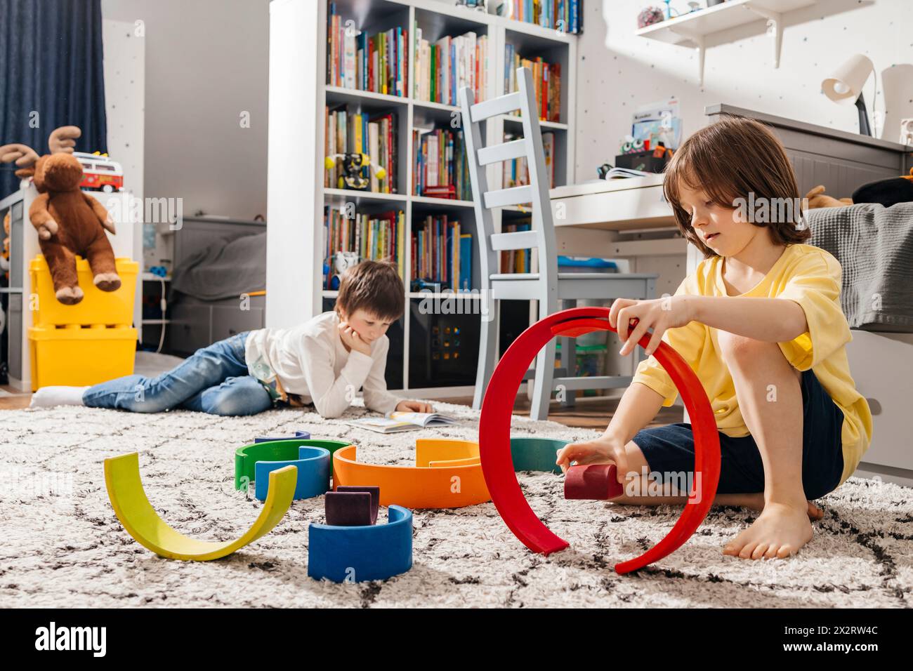 Garçon bâtissant pyramide avec des jouets colorés arc-en-ciel près de frère lisant le livre à la maison Banque D'Images