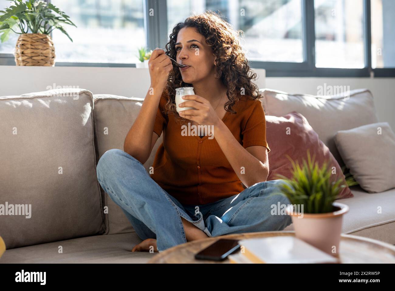 Jeune femme qui mange du yaourt sur un canapé à la maison Banque D'Images