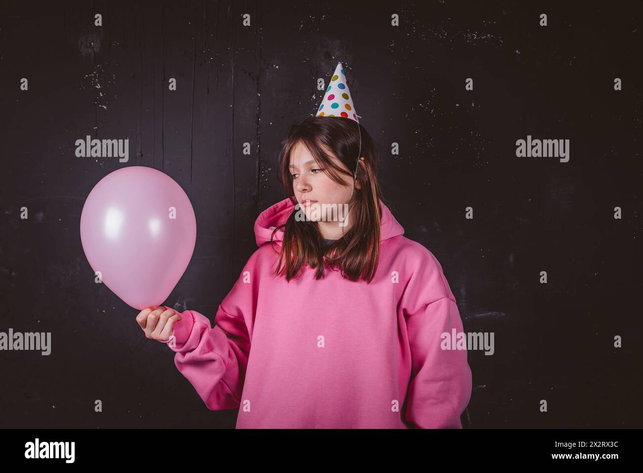 Fille d'anniversaire réfléchie dans le chapeau de fête tenant le ballon rose devant le fond noir Banque D'Images
