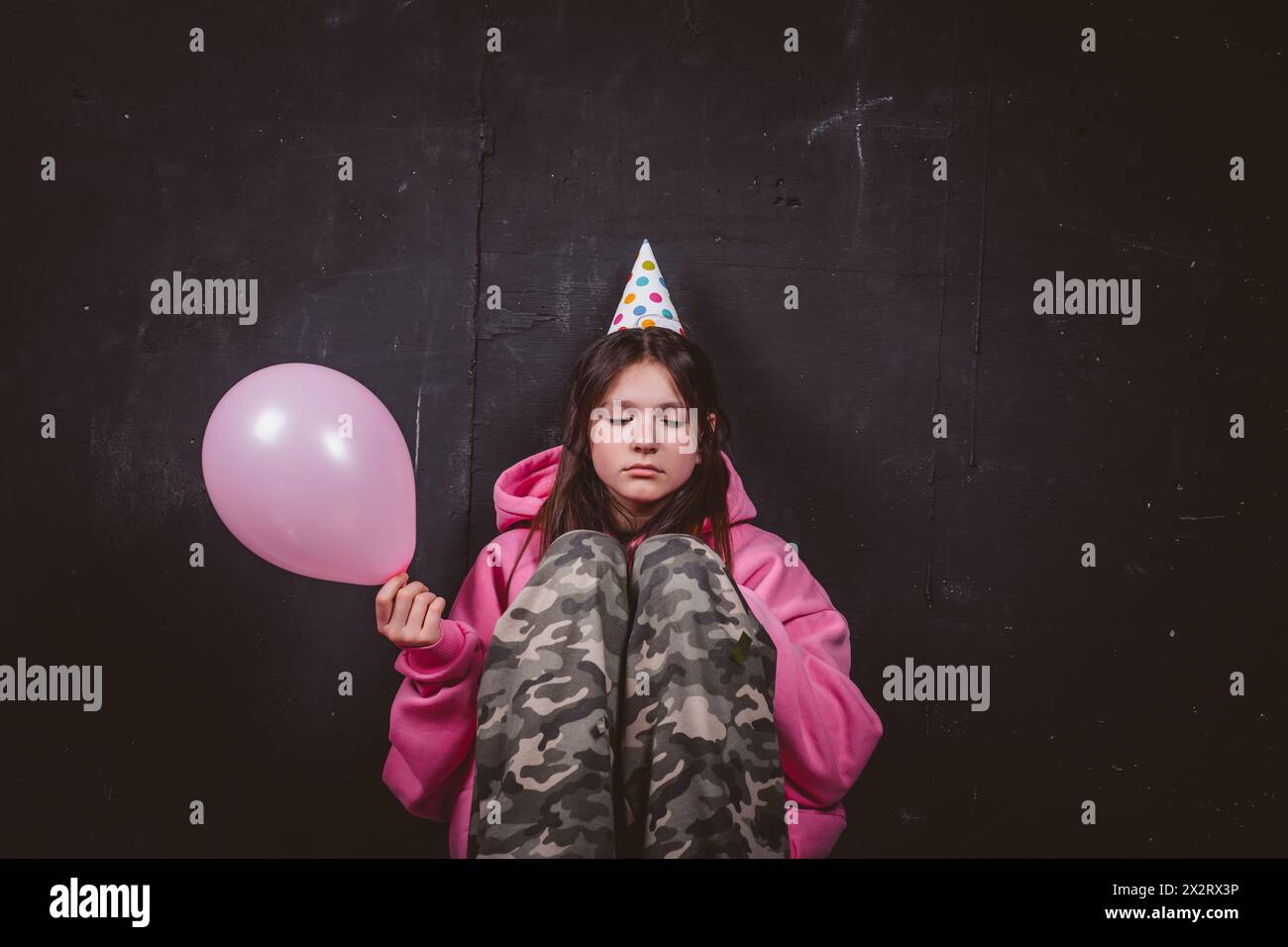 Fille d'anniversaire triste dans le chapeau de fête tenant le ballon rose devant le fond noir Banque D'Images