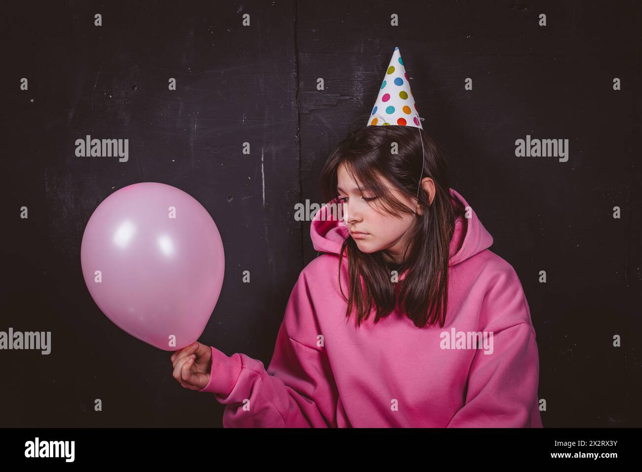 Fille d'anniversaire triste dans le chapeau de fête regardant le ballon rose devant le fond noir Banque D'Images