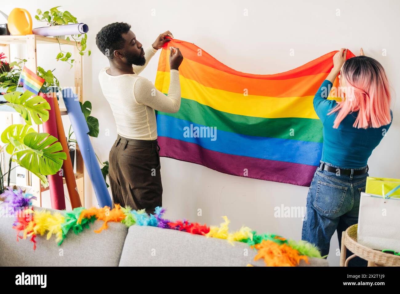 Jeune personne non binaire collant le drapeau arc-en-ciel sur le mur avec un ami à la maison Banque D'Images