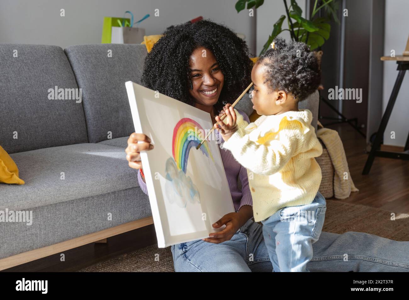 Heureuse mère tenant la toile pour bébé fille peignant arc-en-ciel avec pinceau à la maison Banque D'Images