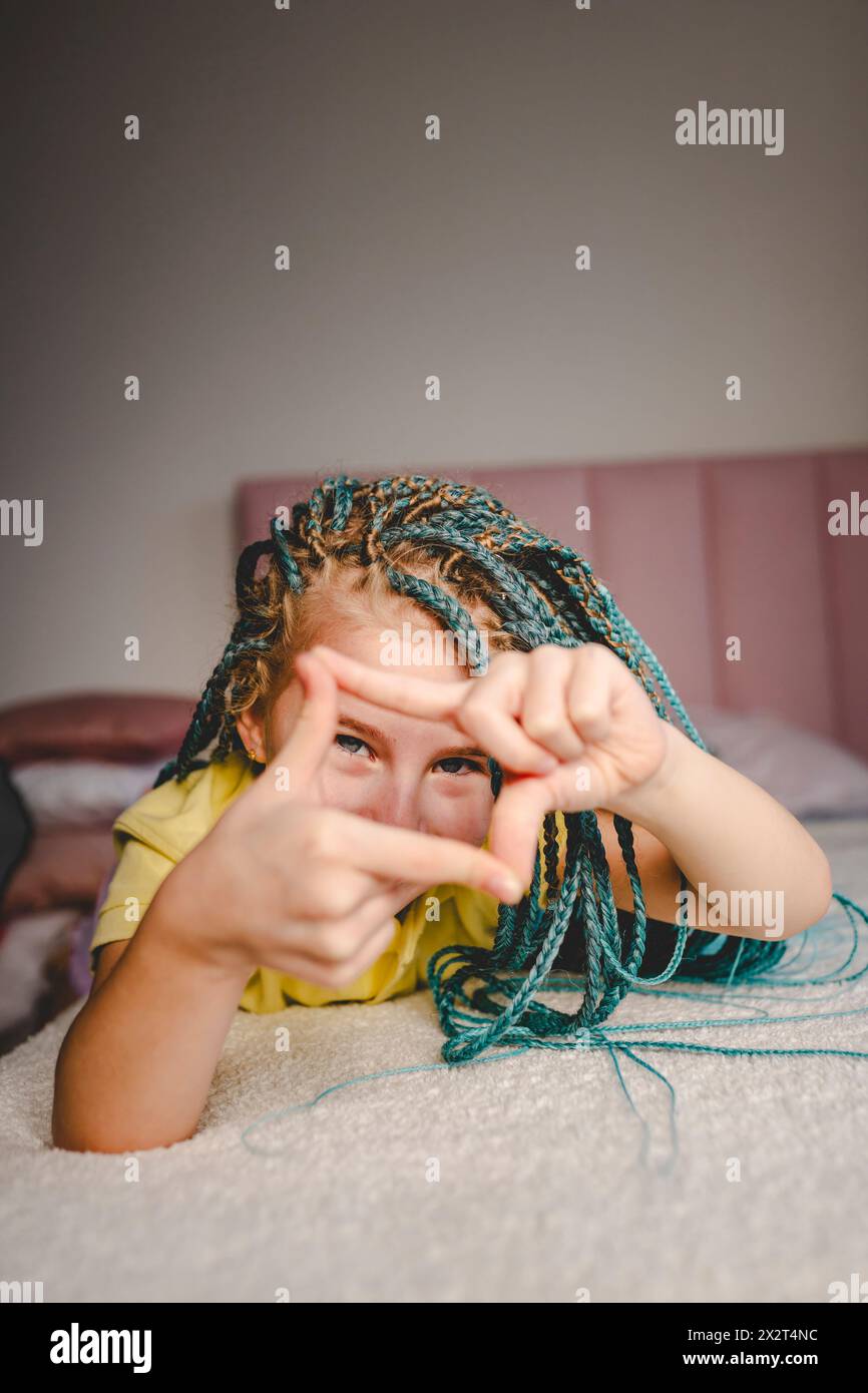 Fille heureuse avec des cheveux tressés teints turquoise couchés sur le lit faisant le cadre de doigt à la maison Banque D'Images