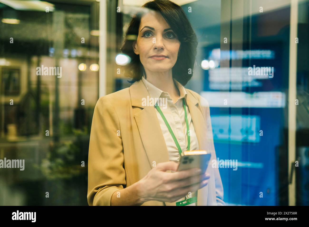Femme d'affaires mature confiante avec téléphone intelligent vu à travers le verre au bureau Banque D'Images
