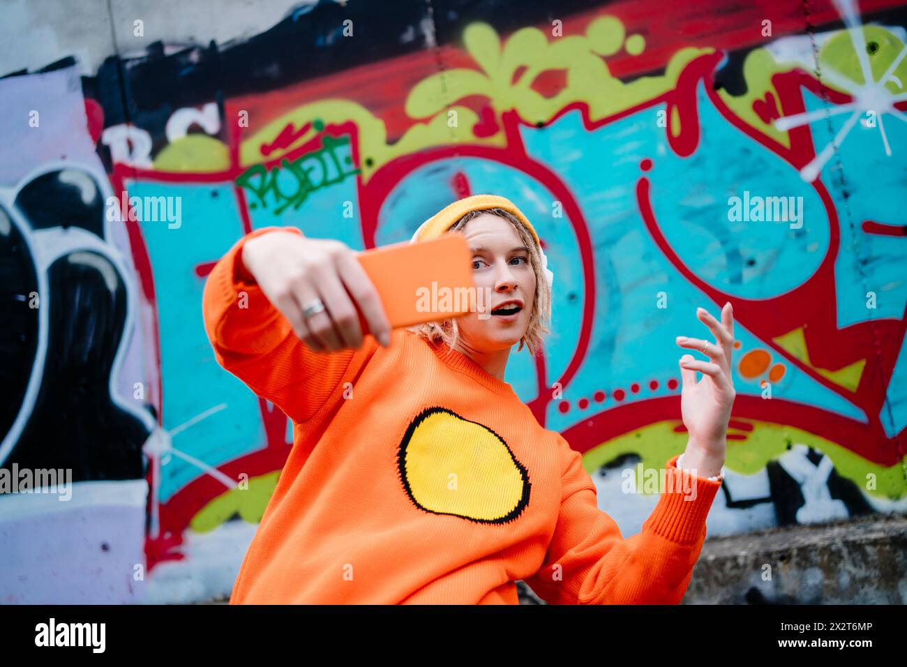 Jeune femme faisant des gestes et prenant selfie à travers un téléphone intelligent devant un mur de graffiti Banque D'Images