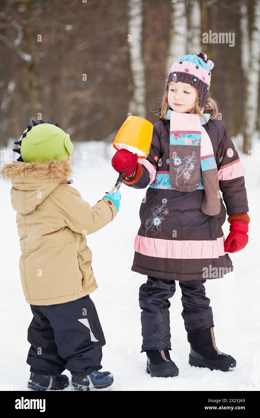 La fille plus âgée donne eskimo fait de neige dans un seau en plastique et une branche cassée à un enfant plus jeune dans le parc d'hiver Banque D'Images