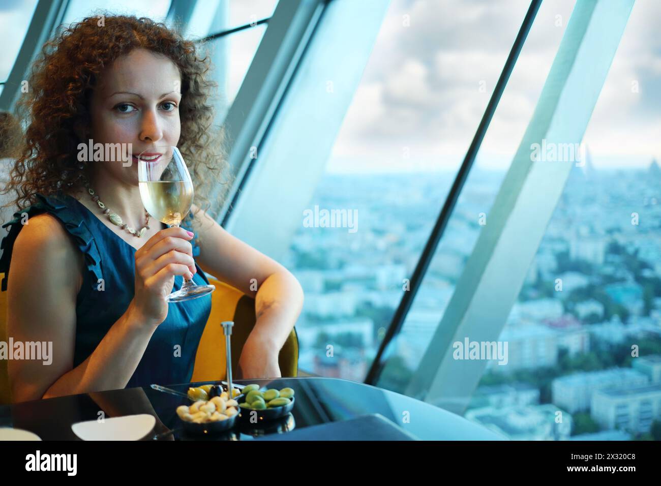 Belle femme boit du vin dans le restaurant, la ville est visible à travers la fenêtre. Banque D'Images