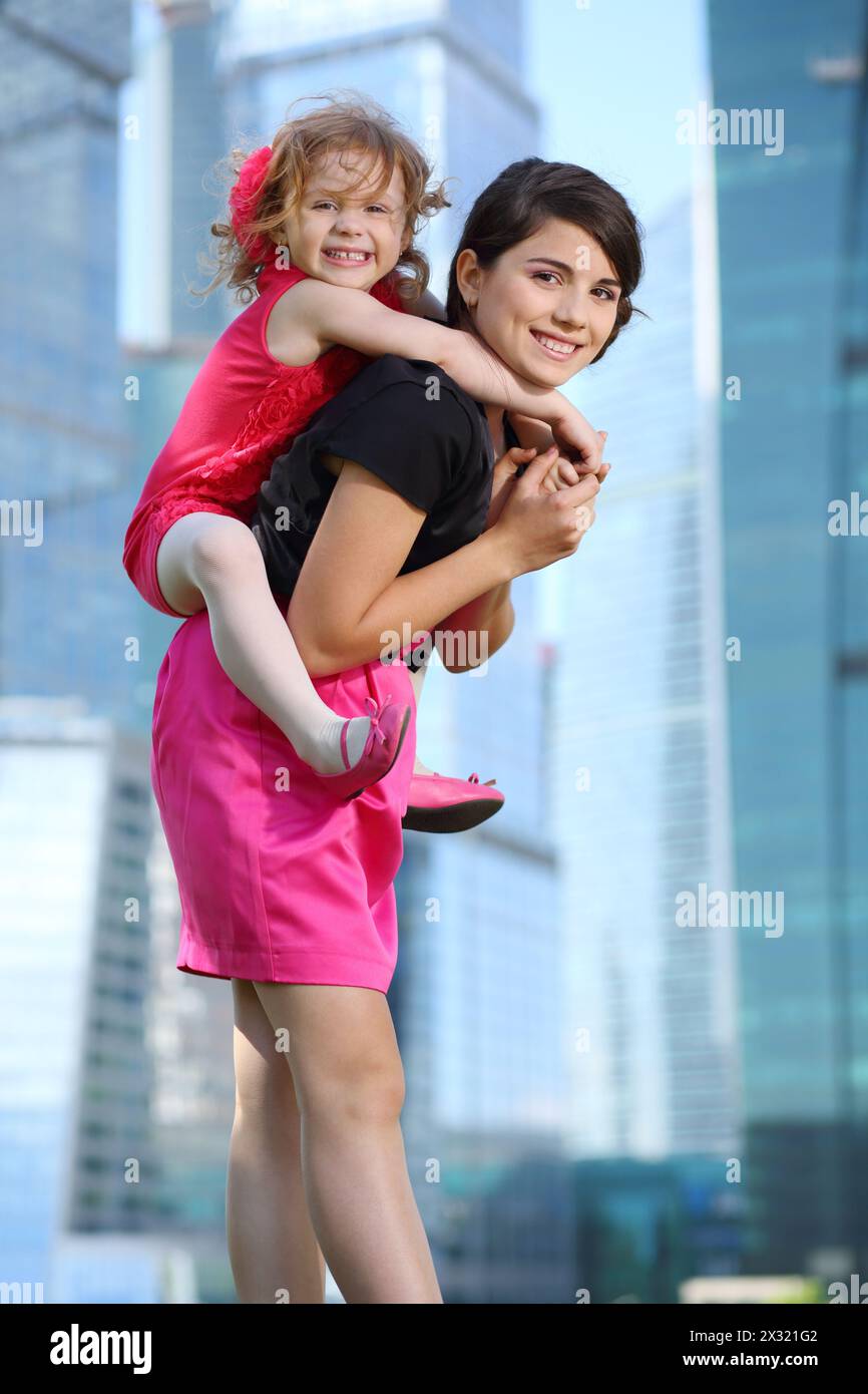 Belle fille porte sur son dos petite fille en rouge près des gratte-ciel bleus. Banque D'Images