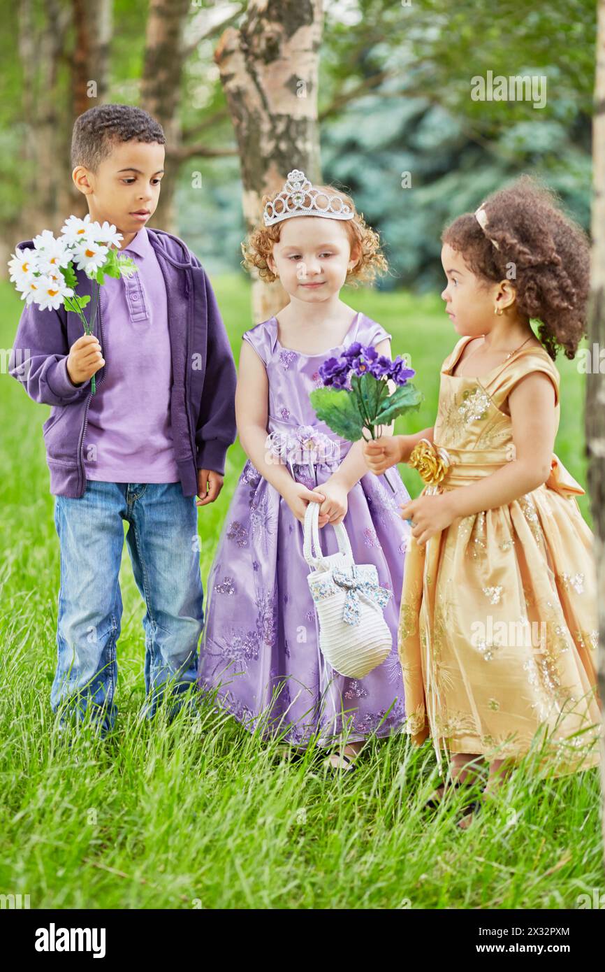 Garçon et deux petites filles habillés dans des blouses de fête se tiennent dans le parc sur la pelouse herbeuse, garçon et une de fille tiennent la fleur Banque D'Images