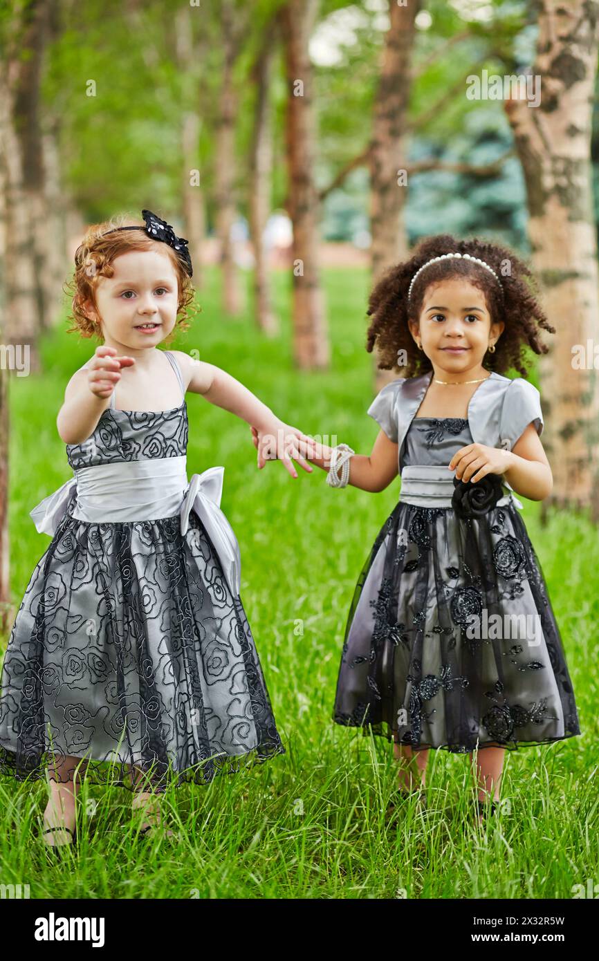 Deux petites filles en robes noires se tiennent debout tenant les mains sur l'herbe entre les arbres dans le parc, se concentrer sur la fille gauche Banque D'Images