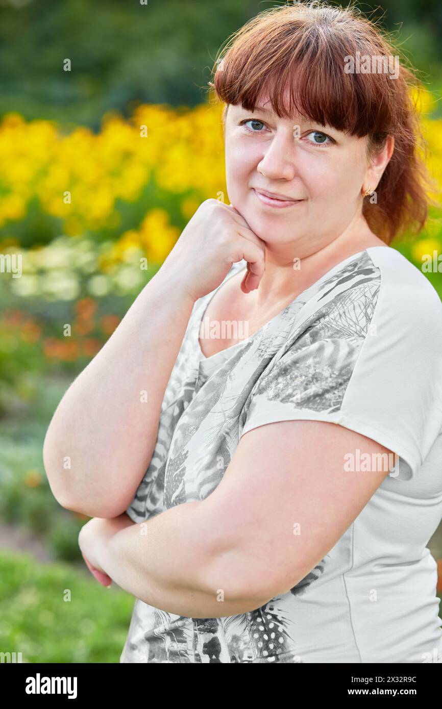 Portrait demi-longueur de femme debout à moitié tournée et soutenant la tête avec la main contre la clairière pleine de fleurs diverses Banque D'Images