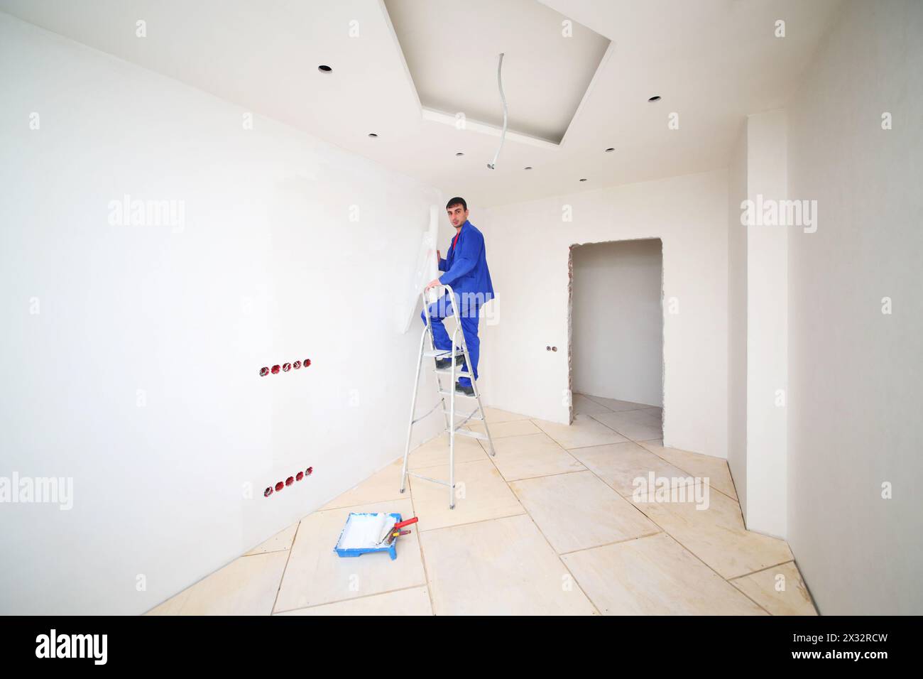 Homme en bleu sur les pâtes d'échelle peignent la fibre de verre dans l'appartement neuf Banque D'Images