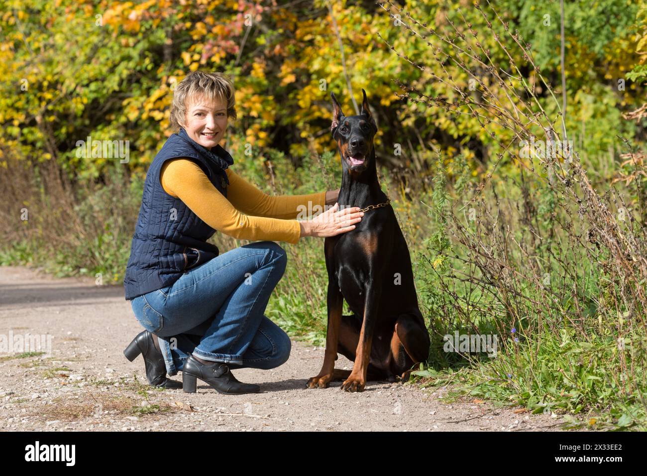 Femme au col de polo beige est assise devant le dobermann dans une forêt. Banque D'Images