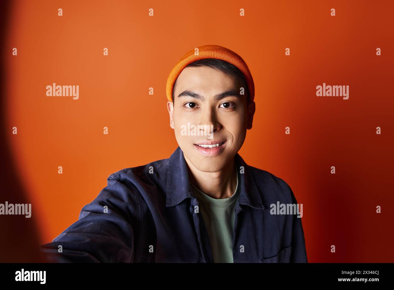 Bel homme asiatique habillé dans une tenue élégante prenant un selfie tout en portant un chapeau orange dans un cadre de studio vibrant. Banque D'Images
