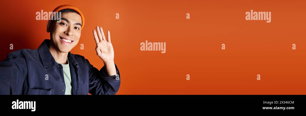 Un homme asiatique élégant dans une veste bleue fait des gestes avec sa main sur un fond orange vif dans un cadre de studio. Banque D'Images