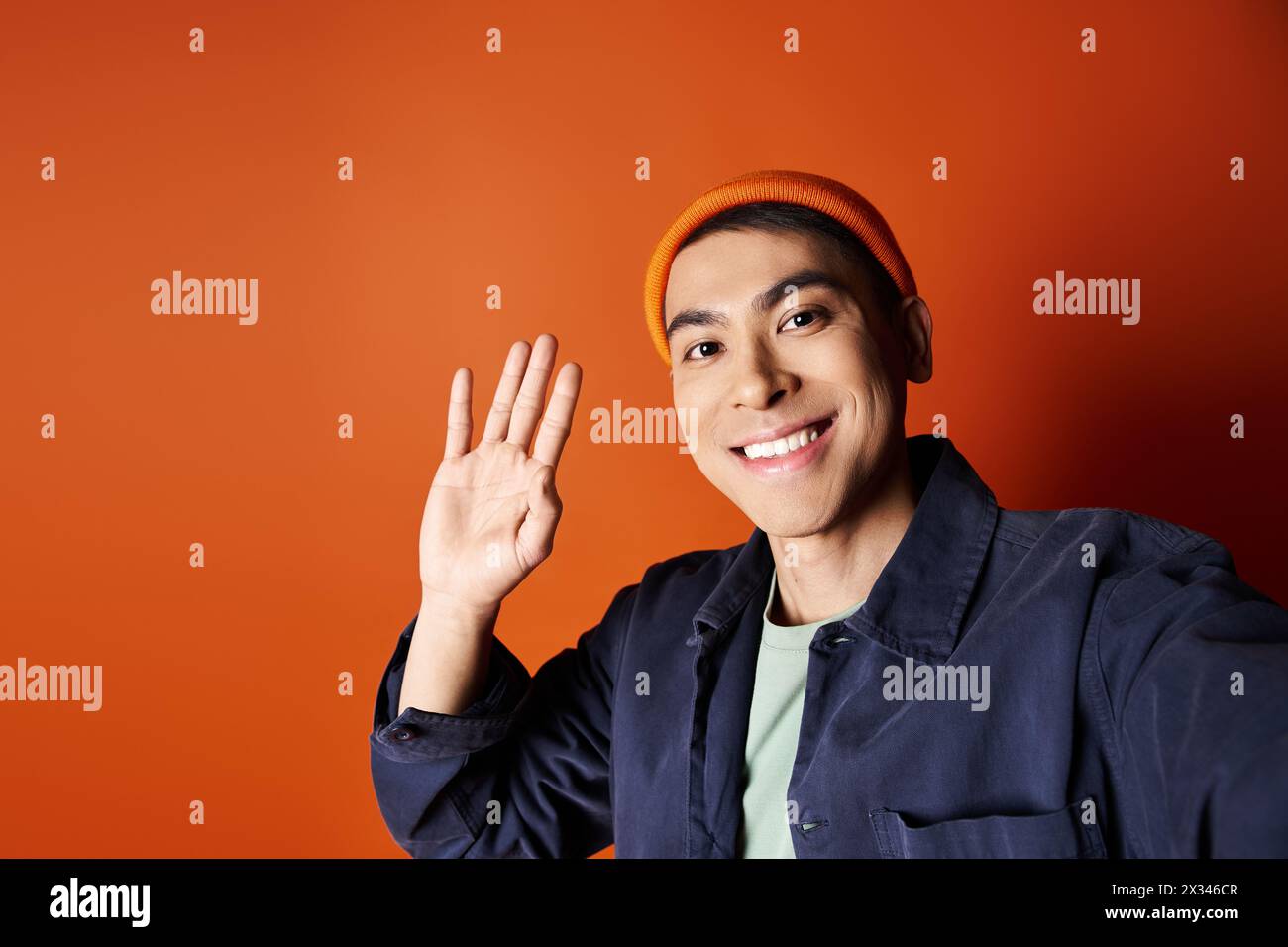 Bel homme asiatique en veste bleue et chapeau orange agitant en toute confiance la main sur un fond orange audacieux. Banque D'Images