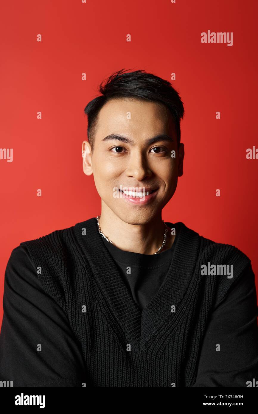 Un bel homme asiatique sourit chaleureusement à la caméra, portant un chandail noir chic sur un fond de studio rouge vif. Banque D'Images