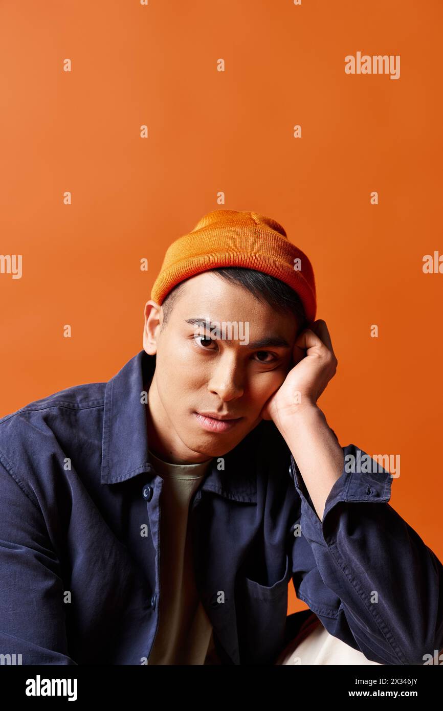 Bel homme asiatique vêtu d'une chemise bleue et d'un chapeau orange, debout avec confiance sur un fond orange vif dans un studio. Banque D'Images