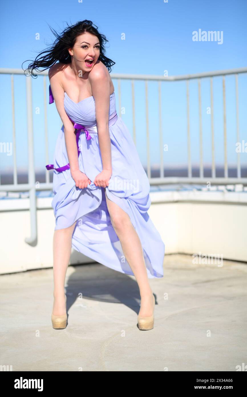 Jolie fille en robe se tient sur le toit d'un grand bâtiment et crie sur le vent à la journée ensoleillée Banque D'Images