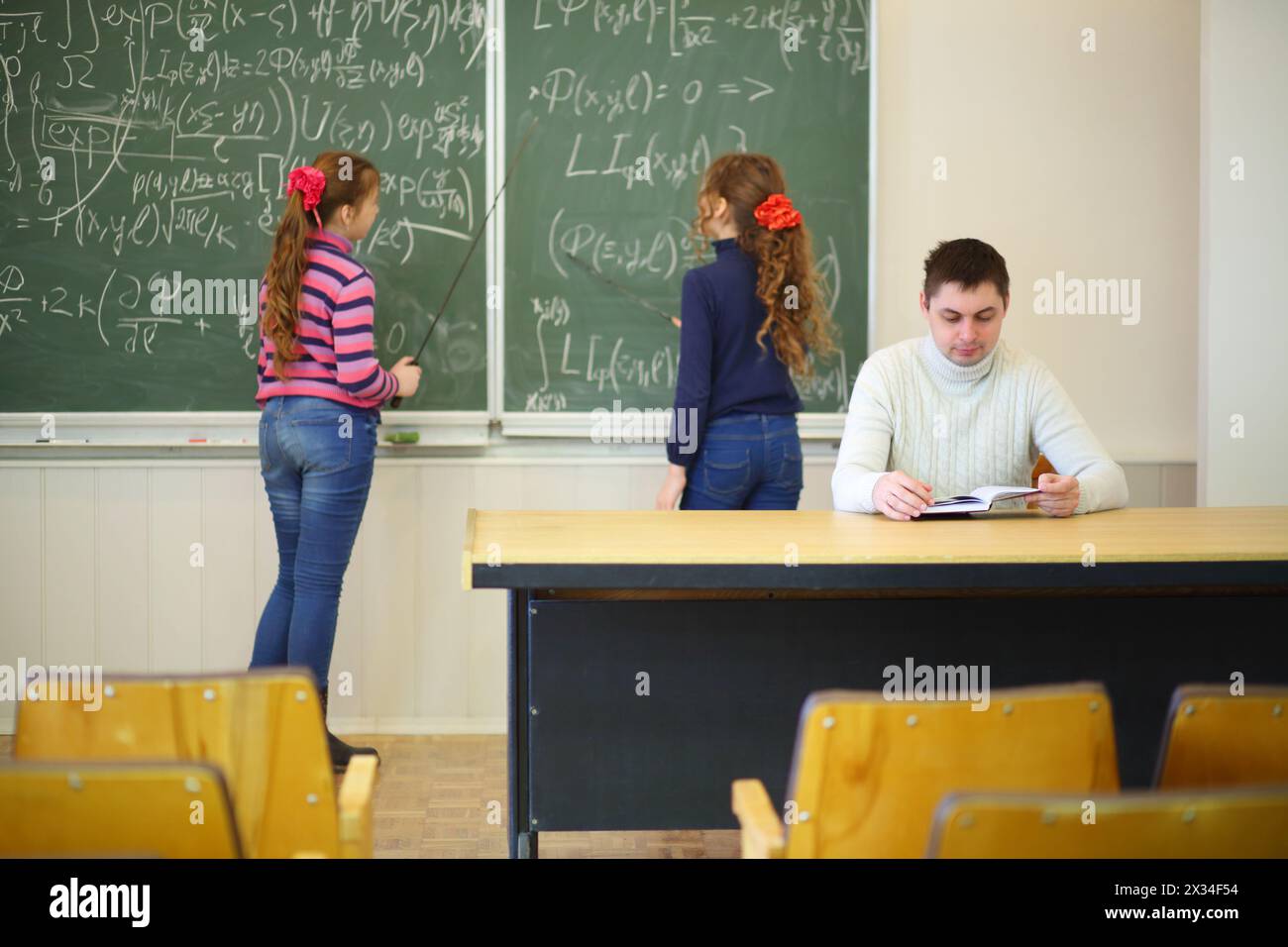 Deux filles avec des pointeurs se tiennent près du tableau noir avec des formules et le professeur regarde dans le livre en classe, se concentrer sur l'homme Banque D'Images