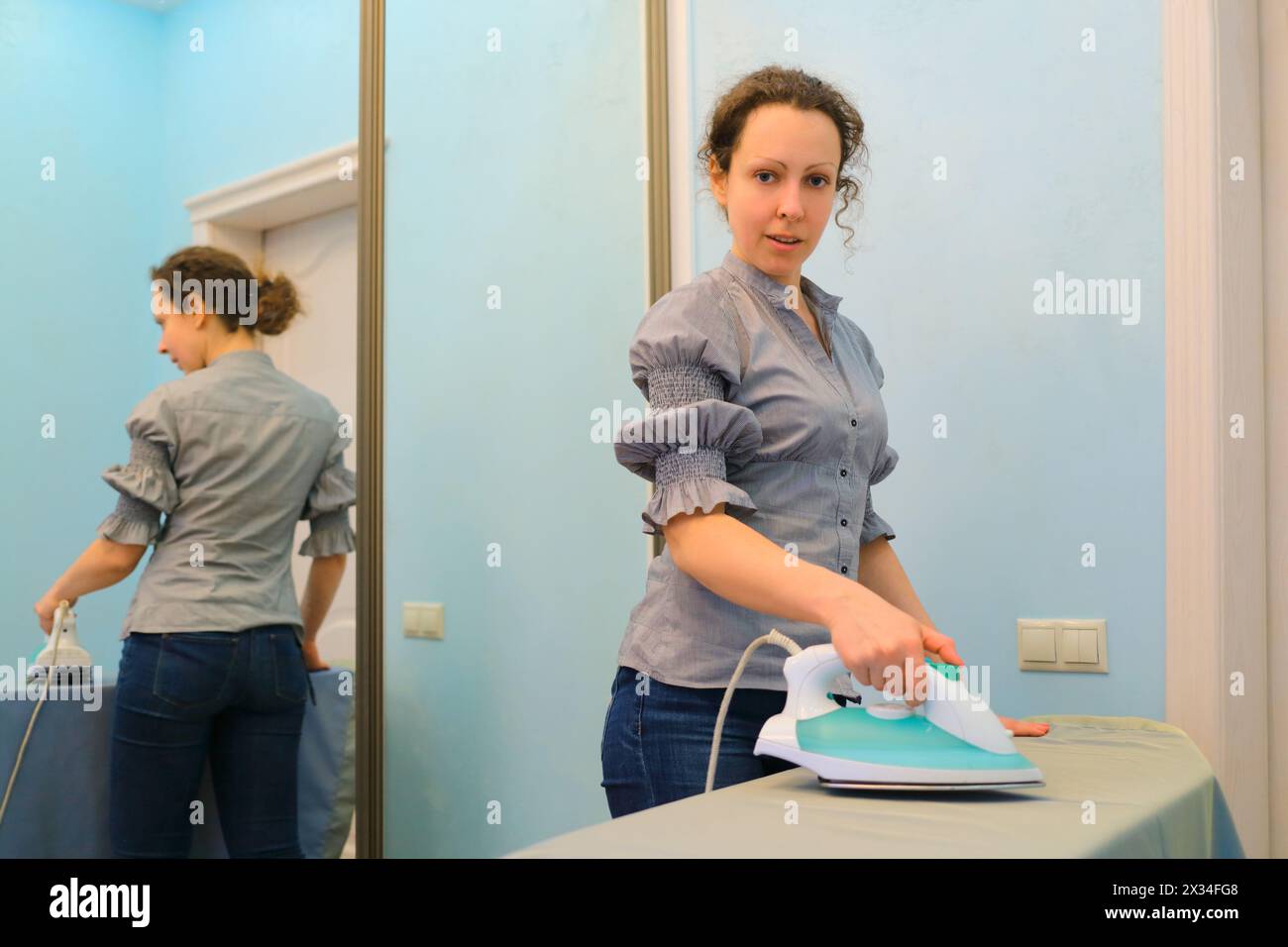 jeune femme au foyer en blouse grise et jeans repassait dans la chambre, reflétée dans le miroir Banque D'Images