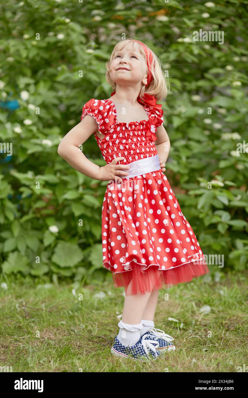 Petite fille en robe à pois rouges se tient avec ses mains akimbo et regardant vers le haut à la pelouse herbeuse. Banque D'Images