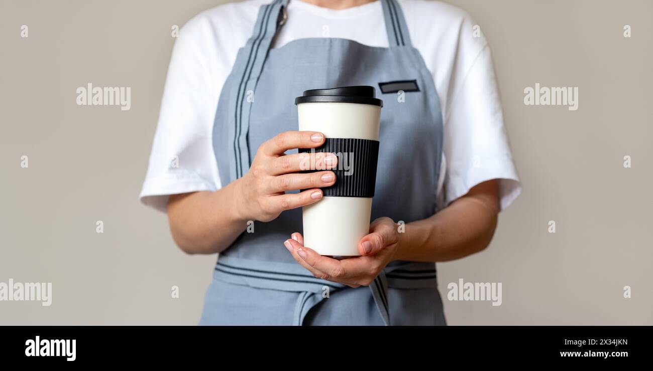Une barista portant un tablier tient une grande tasse de café réutilisable devant elle. Banque D'Images