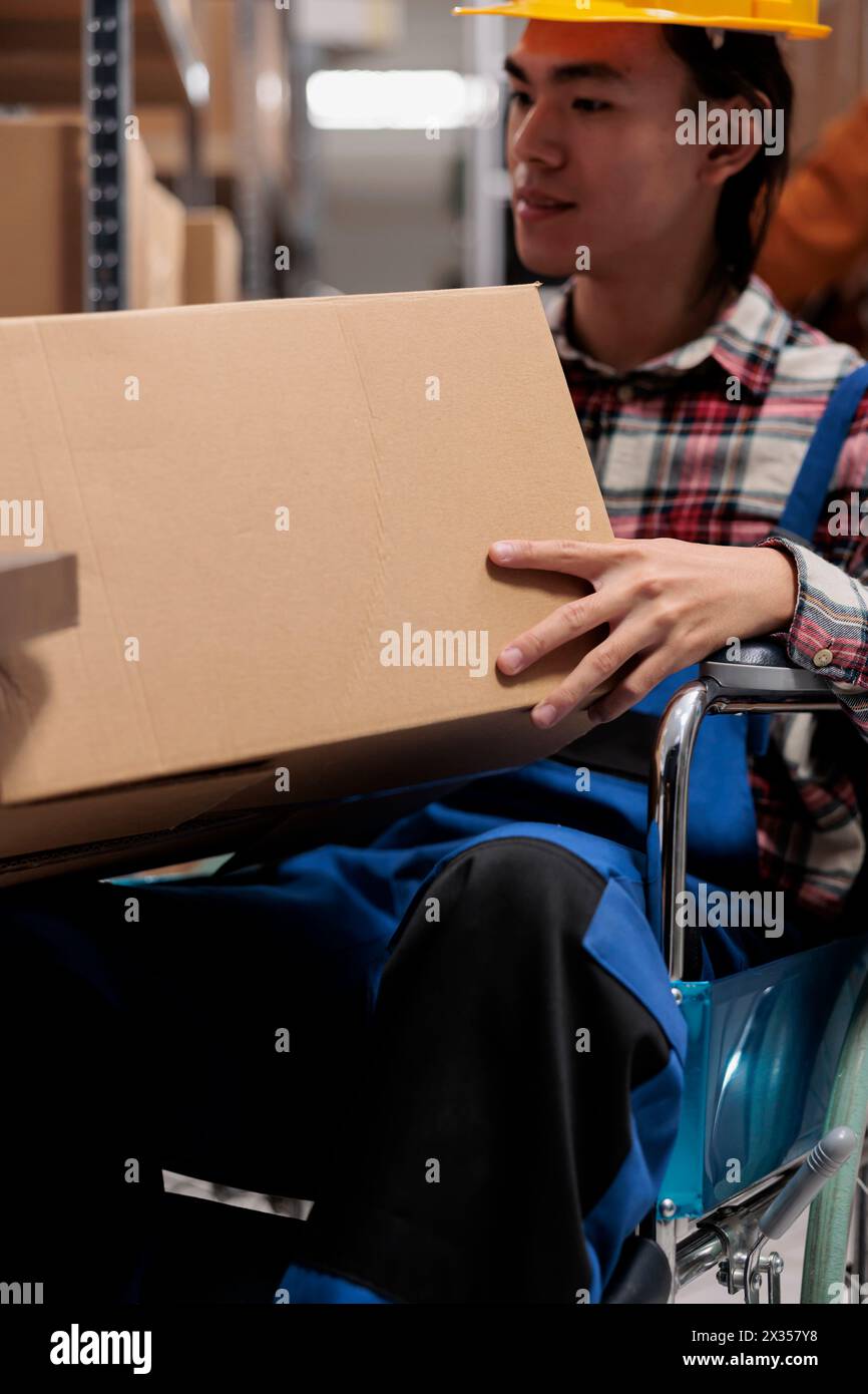 Utilisateur de fauteuil roulant de préparateur de commande d'entrepôt asiatique tenant le colis du client. Jeune employé d'entrepôt handicapé préparant le fret pour l'expédition tout en travaillant dans un lieu de travail inclusif Banque D'Images
