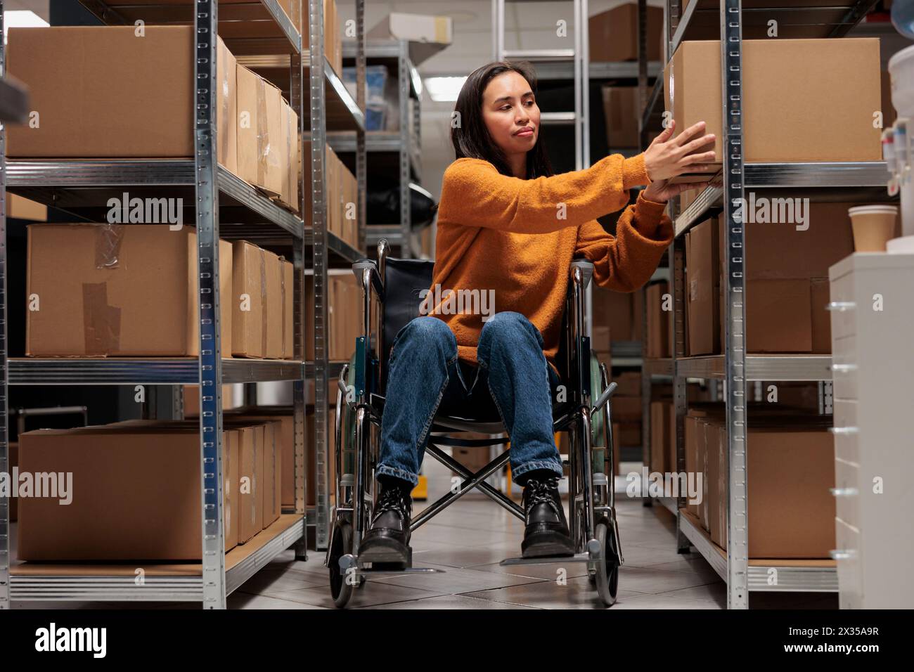 Femme utilisateur de fauteuil roulant prenant la boîte en carton de l'étagère pour préparer la commande du client pour l'expédition dans l'entrepôt. Manipulateur de colis asiatique tenant le colis, travaillant dans un entrepôt favorable aux personnes handicapées Banque D'Images