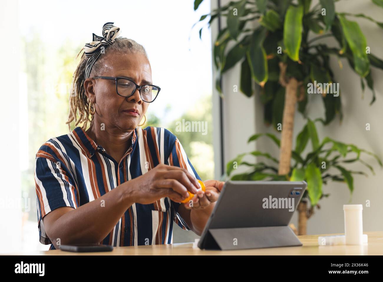 Une femme afro-américaine senior portant des lunettes utilise une tablette à la maison et prend des médicaments, copie l'espace. Elle a les cheveux gris attachés, et porte un Banque D'Images