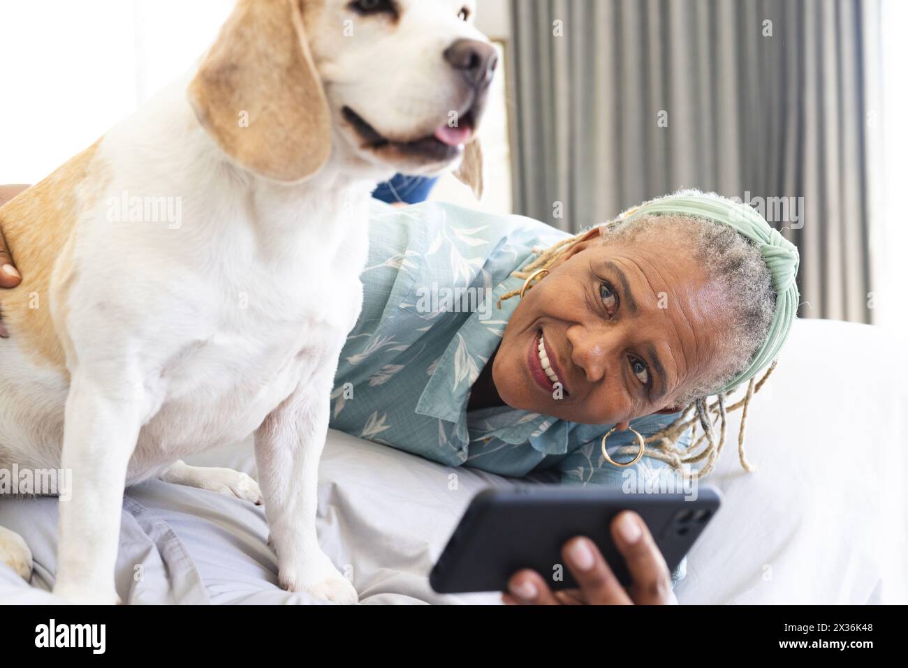 Une femme afro-américaine sénior est allongée sur le lit à la maison, tenant un smartphone, prenant un selfie avec son chien. Elle a les cheveux gris, porte un bleu clair Banque D'Images