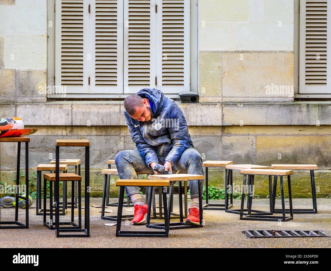 Homme sans masque de protection à l'aide d'un outil électrique ponçant les dessus en bois des tabourets - Tours, Indre-et-Loire (37), France. Banque D'Images