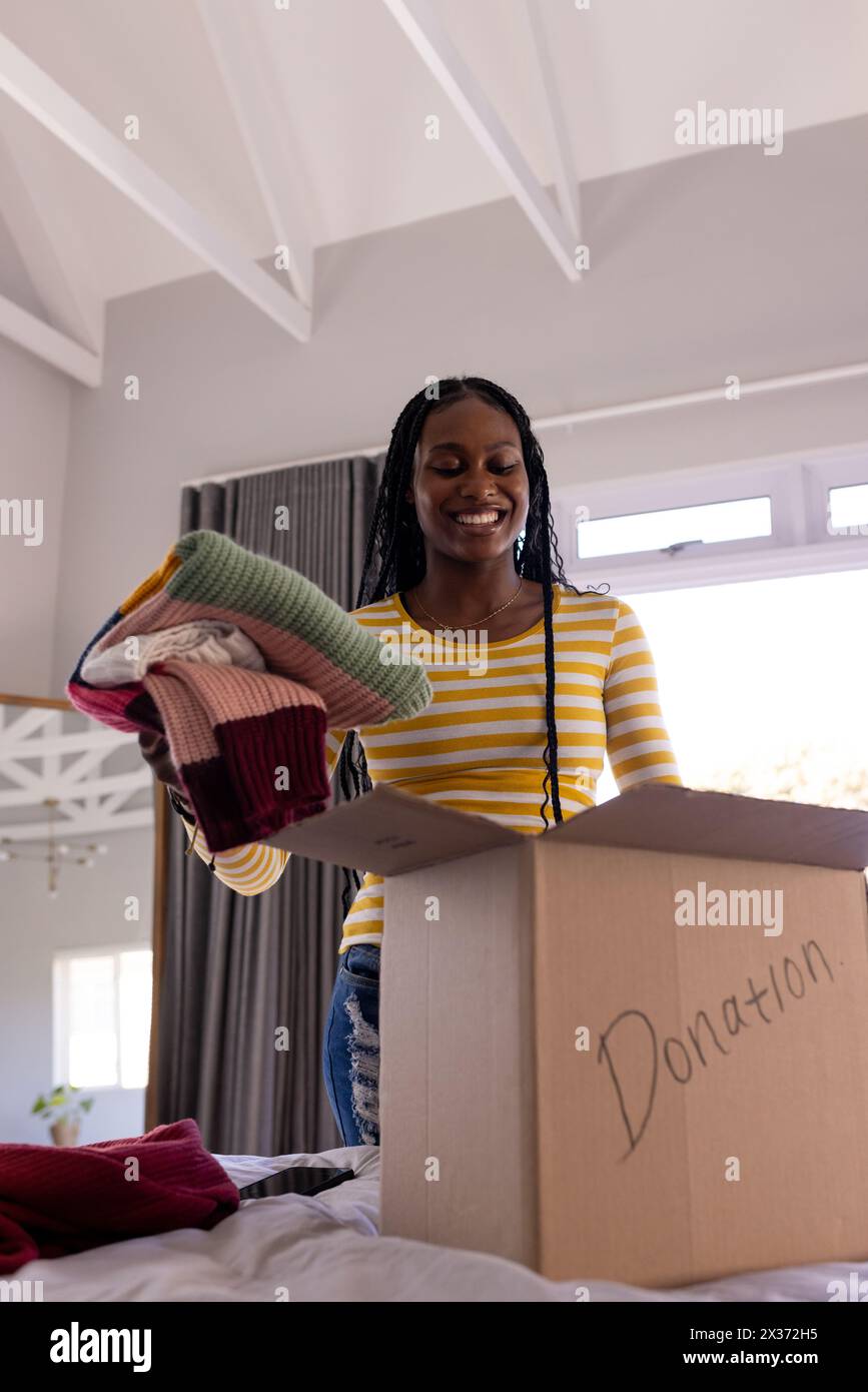 Une jeune femme afro-américaine est à la maison, emballant des vêtements dans une boîte de dons Banque D'Images