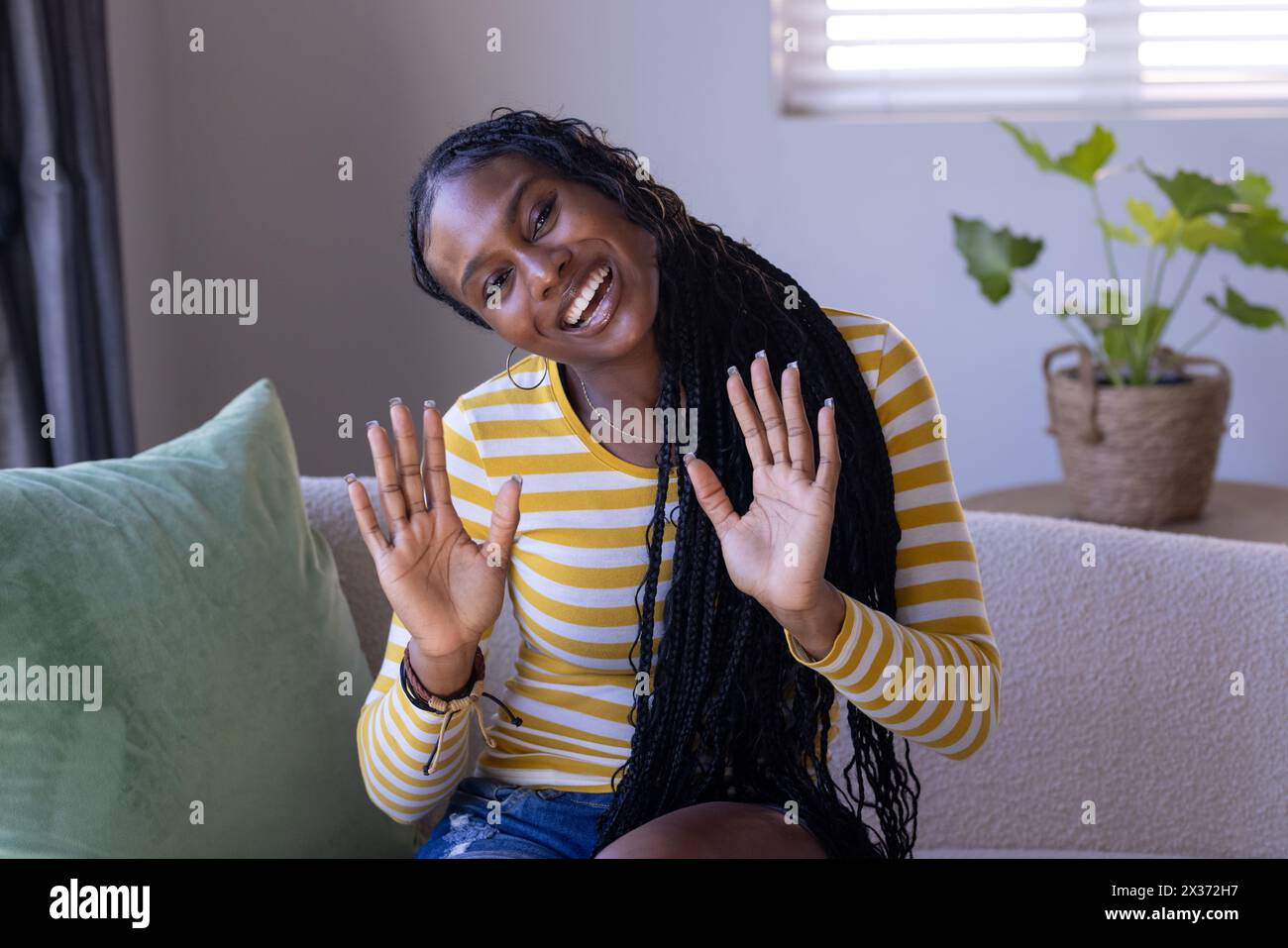 Une jeune femme afro-américaine assise à la maison, agitant les mains avec un sourire éclatant pendant un appel vidéo. Portant une chemise rayée jaune, longue tressée ha Banque D'Images