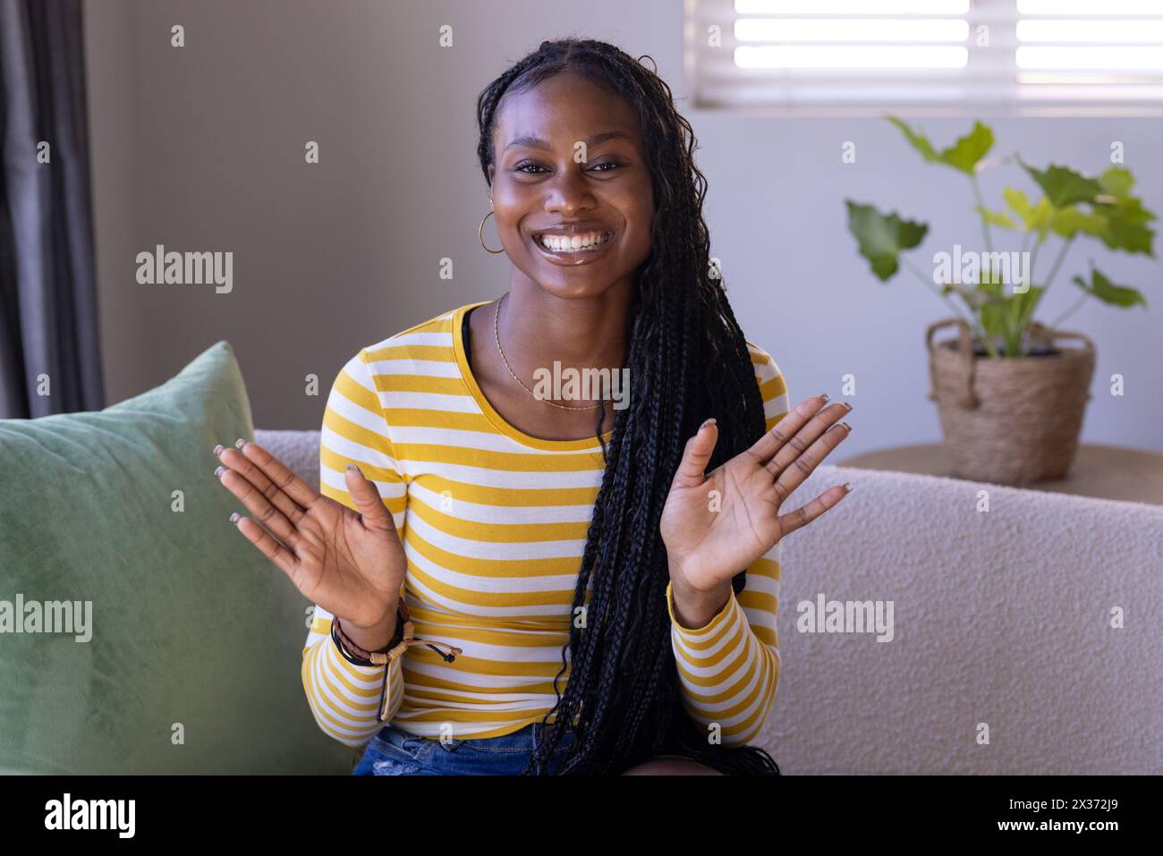 Jeune femme afro-américaine assise à la maison, applaudissant des mains, portant une chemise rayée pendant un appel vidéo, espace copie. Elle a de longs cheveux tressés, un frère Banque D'Images