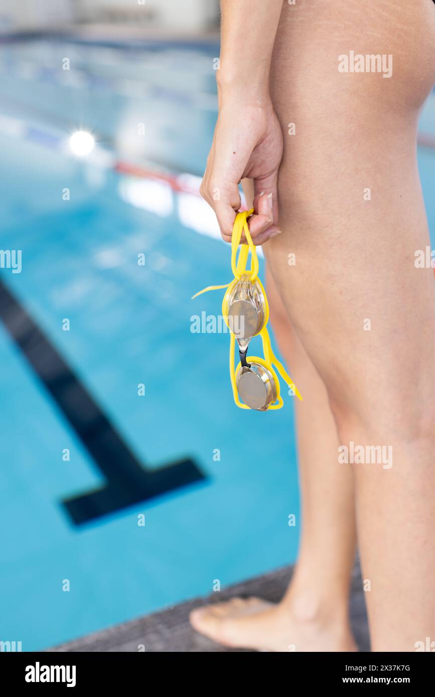 Jeune femme nageuse biraciale tenant des lunettes jaunes se tient près de la piscine à l'intérieur, espace de copie Banque D'Images