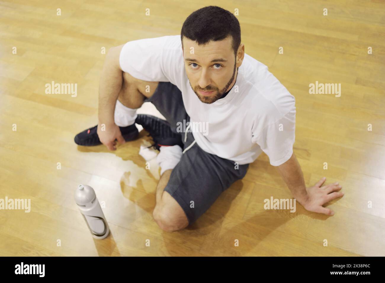 Bel homme est assis sur le sol avec une bouteille de cocktail sportif dans la salle de gym après l'entraînement Banque D'Images