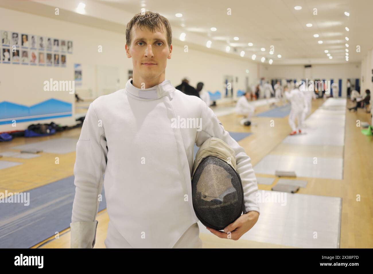 Homme en costume avec des stands de casque dans le gymnase pour la formation d'escrime, d'autres personnes hors de portée Banque D'Images