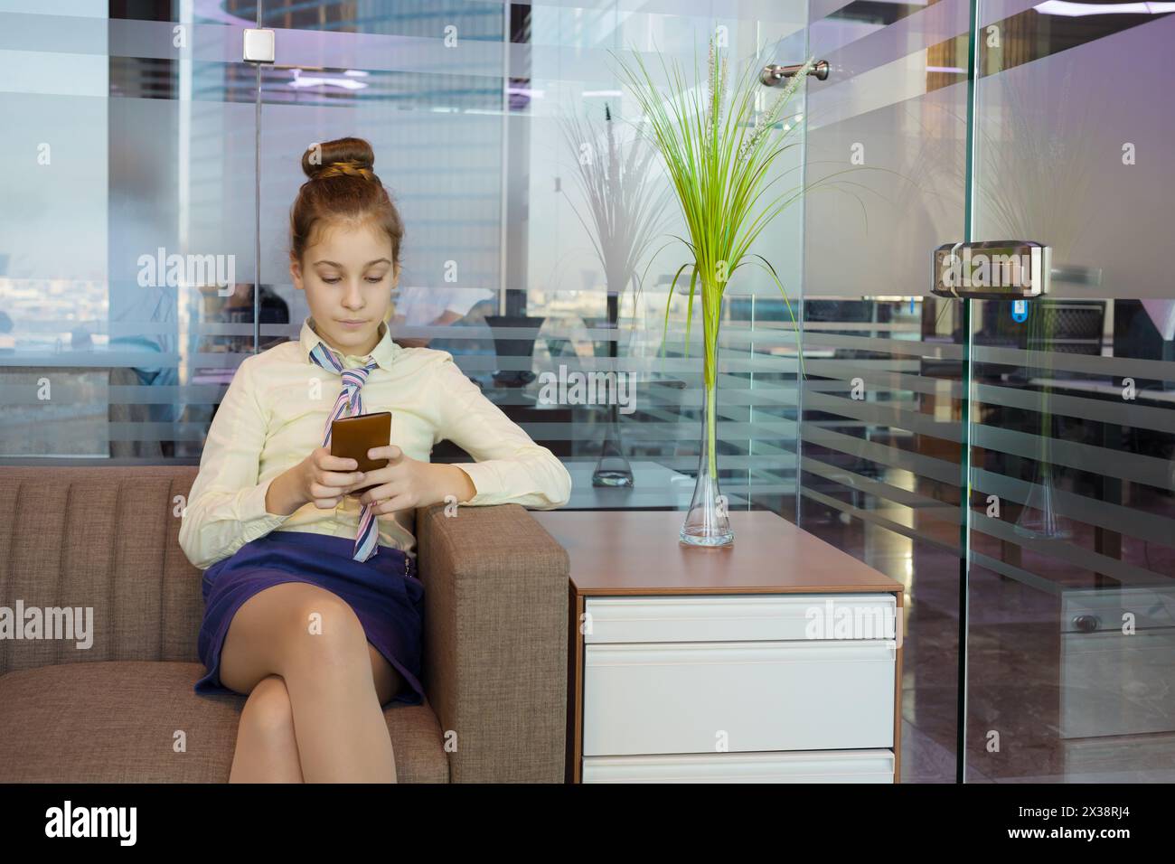 Jolie fille est assise sur un canapé avec téléphone portable dans un bureau moderne avec des murs de verre Banque D'Images